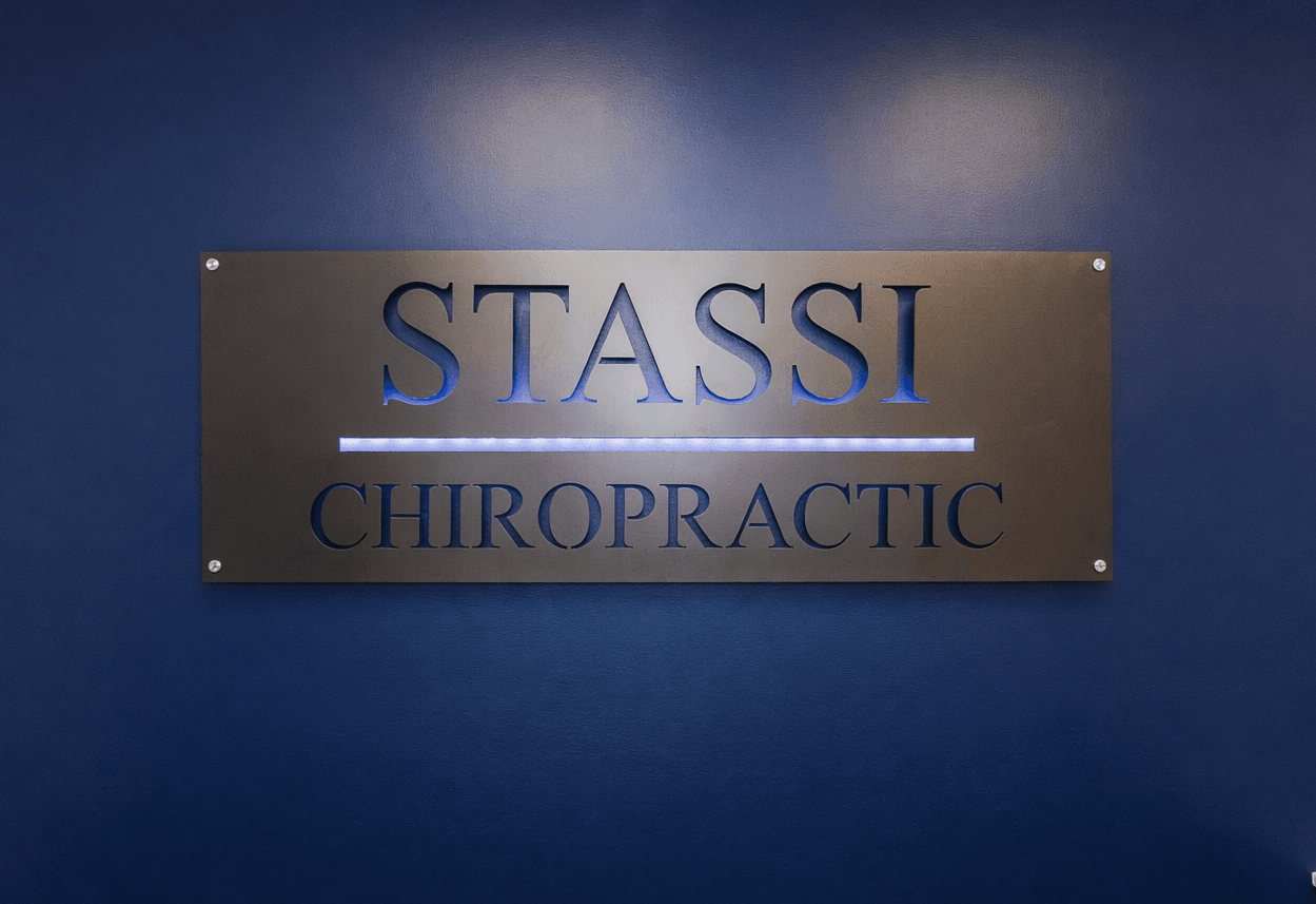 Five smiling Stassi Chiropractic team members, four women and one man, pose in their modern office, with the man flexing a bicep.
