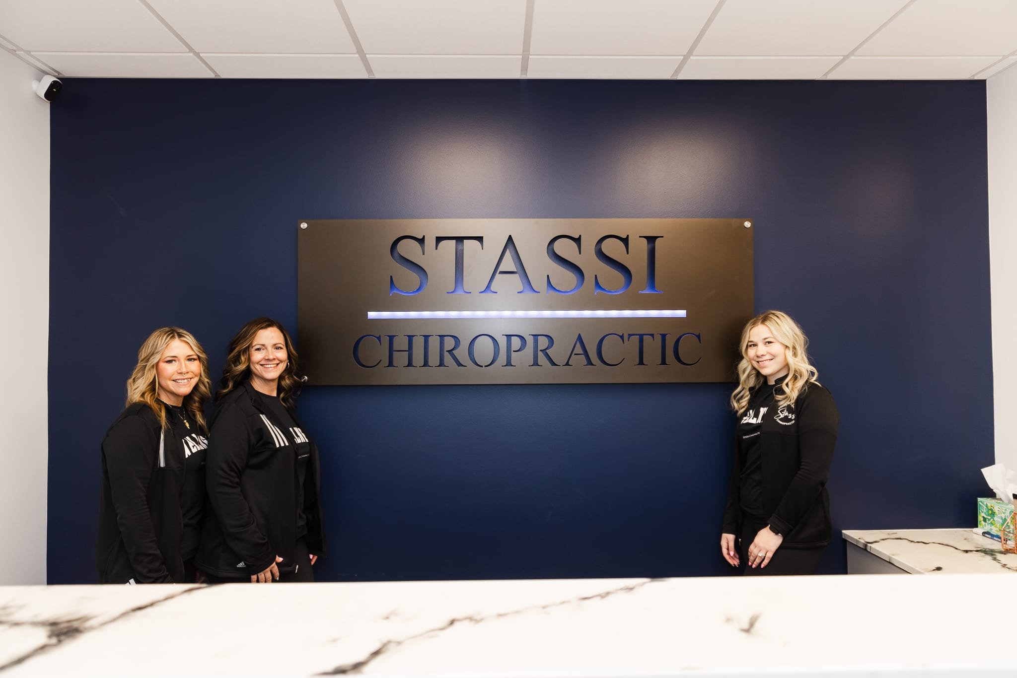 Three smiling team members of Stassi Chiropractic, dressed in black branded shirts, pose for a professional photo.